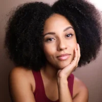 A woman with natural curly hair, wearing a sleeveless burgundy top, rests her face on her hand and looks at the camera with a neutral expression.