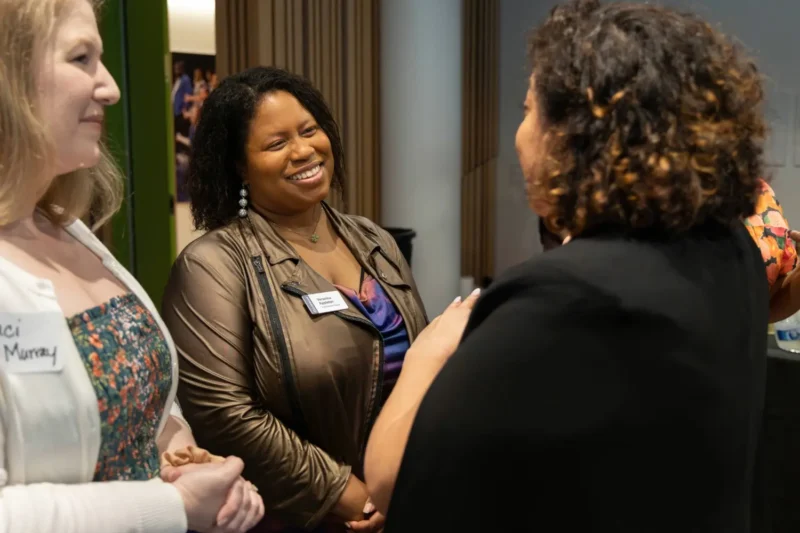 Three women stand together indoors, engaged in conversation and smiling. Two wear name tags and business casual attire.