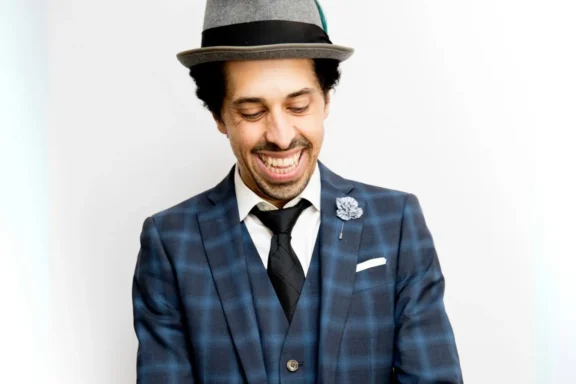 A man in a blue plaid suit, black tie, and gray hat smiles while looking down. He has a boutonnière on his lapel and stands against a plain white background.