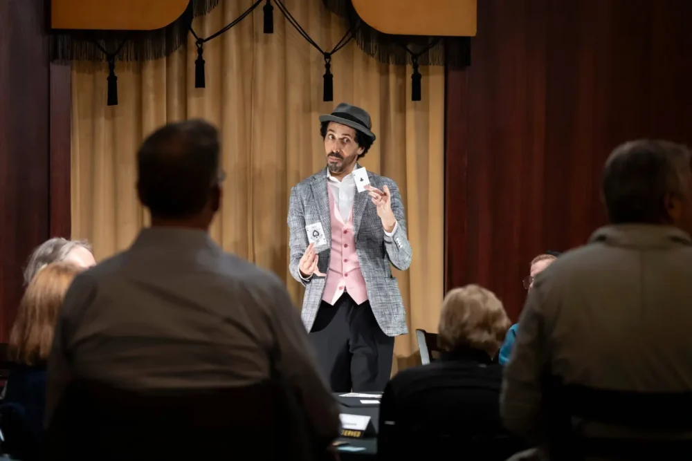 A magician in a suit, vest, and hat performs a card trick on stage in front of an audience seated in a dimly lit room.