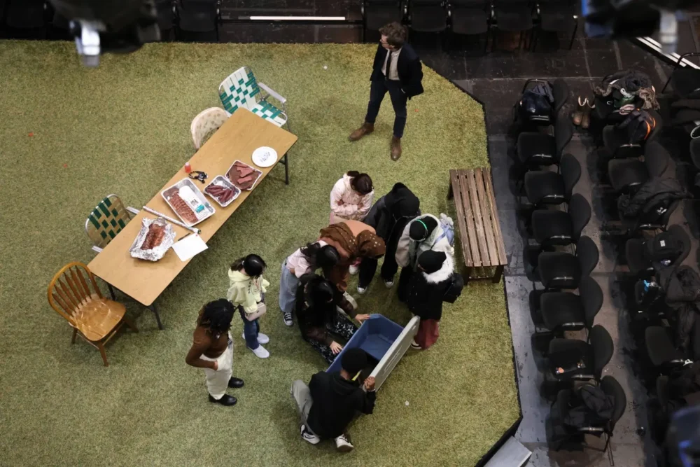 A group of people gather around a blue container on a grassy stage near a table with food, while one person stands apart. Empty chairs are arranged along the edge.