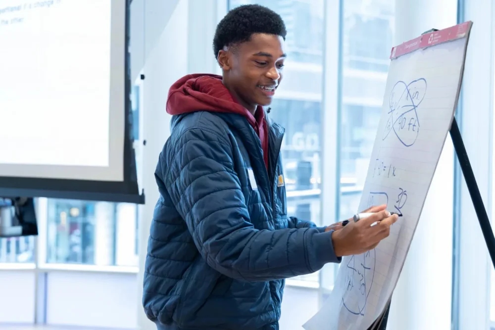 A person in a blue jacket draws diagrams with a marker on a flip chart during a presentation in a bright, modern room.