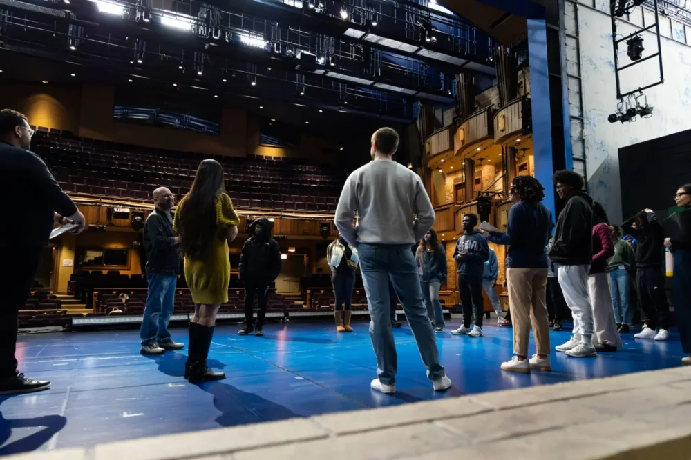 A group of people stand in a circle on a theater stage, with empty audience seats and lighting equipment visible in the background.
