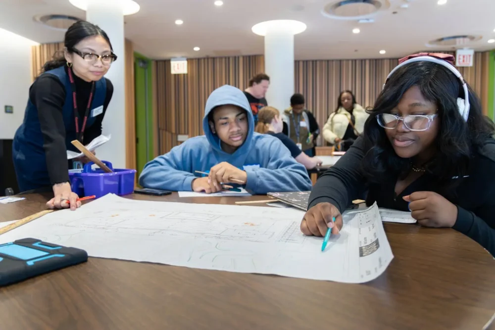 Three students sit at a table examining a large sheet of paper with drawings, while an instructor observes and other people are visible in the background.