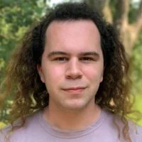 A person with long, curly brown hair and a light complexion is standing outdoors, facing the camera, with trees and greenery in the background.
