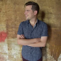 A man with short hair stands against a textured, brownish brick wall with his arms crossed, smiling and looking to his left.