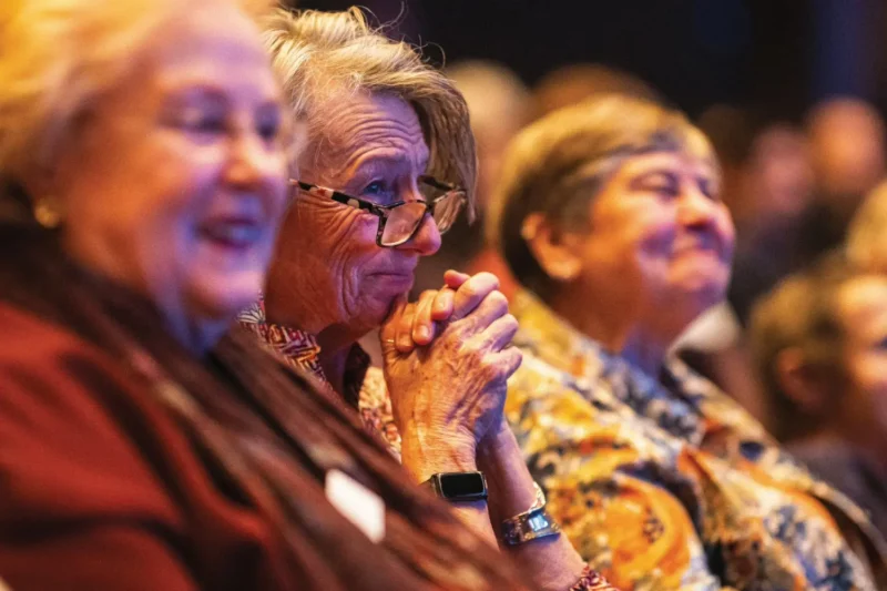 A group of older women sit closely together in an audience, appearing engaged and attentive, with one woman clasping her hands and wearing glasses.