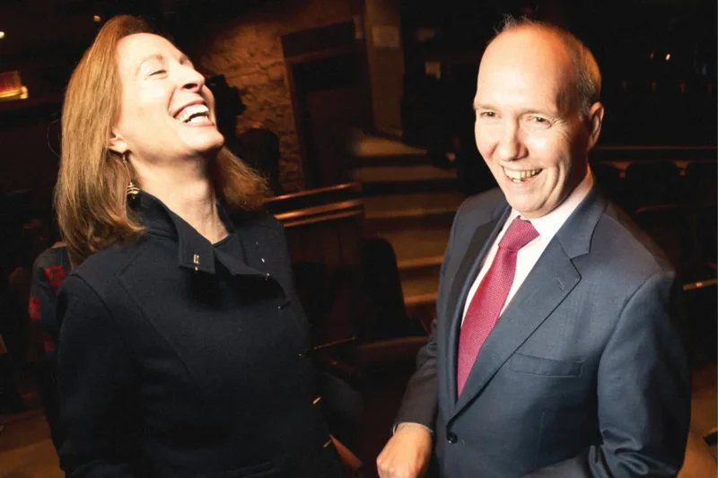 Two adults in formal attire stand indoors, smiling and laughing together in a dimly lit setting.
