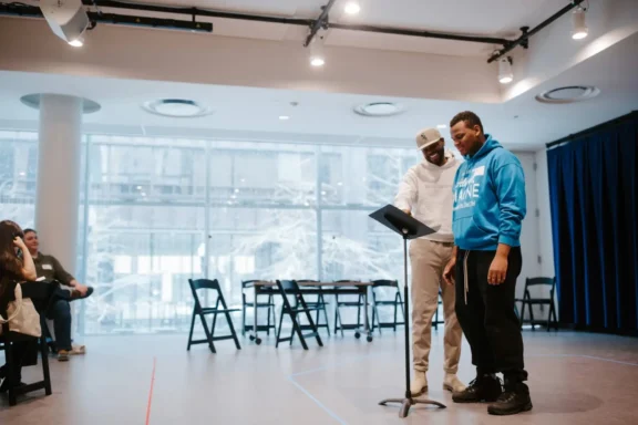 Two men stand at a music stand in a bright room with large windows, reading or rehearsing, while seated people observe in the background.
