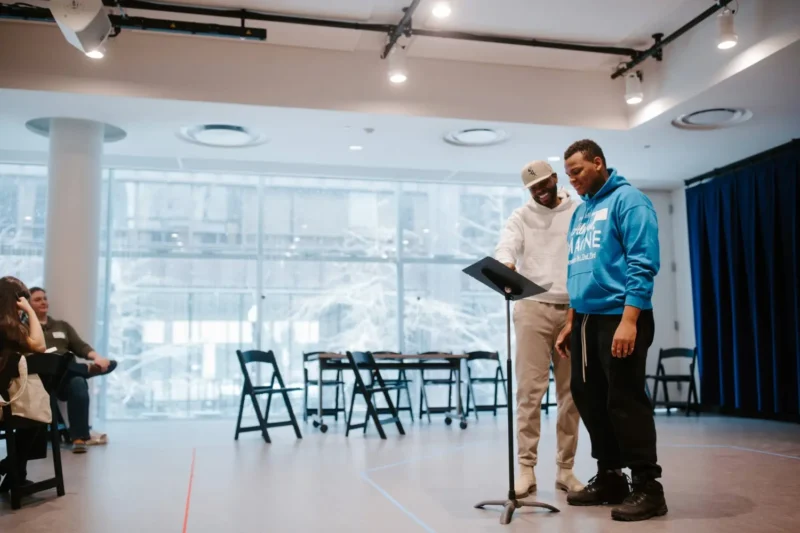 Two men stand at a music stand in a bright room with large windows, reading or rehearsing, while seated people observe in the background.