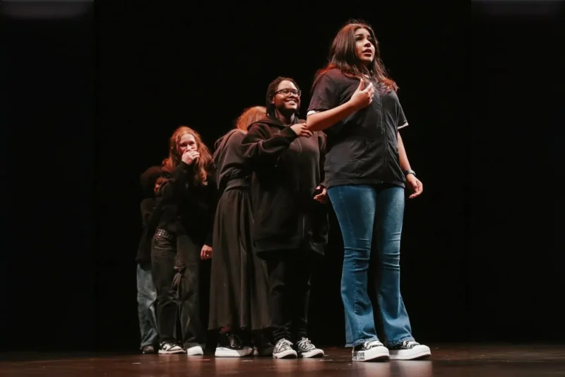 Five teenagers stand on stage in a line, with the person in front speaking and the others behind her, all wearing dark clothing and sneakers in a theater setting.