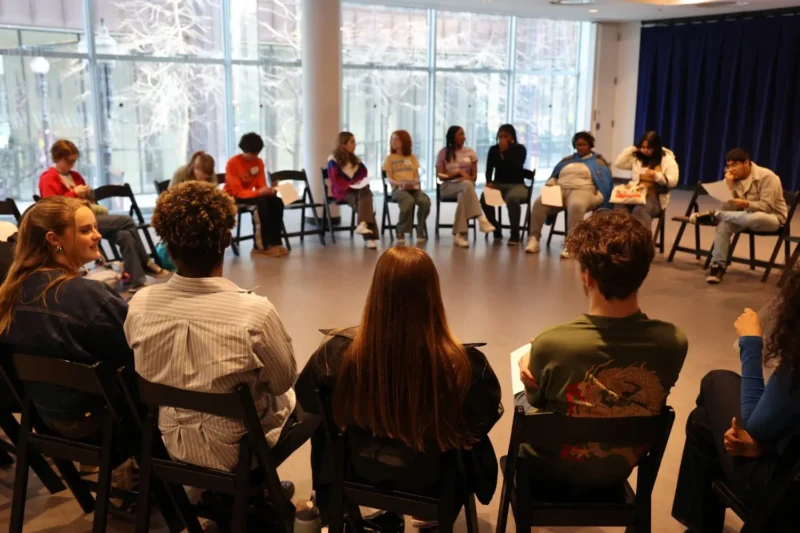 A group of people sit in a large circle of chairs in a bright room, engaged in discussion or an activity.