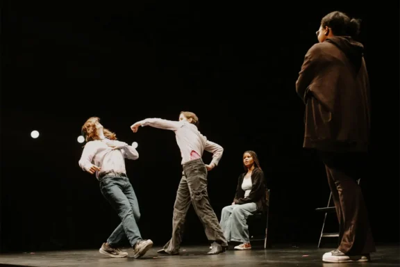 Four people are on a dark stage. One person throws a punch at another who leans back. A third person sits on a chair, while a fourth stands in the foreground, watching.