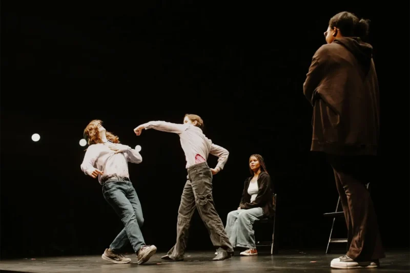 Four people are on a dark stage. One person throws a punch at another who leans back. A third person sits on a chair, while a fourth stands in the foreground, watching.