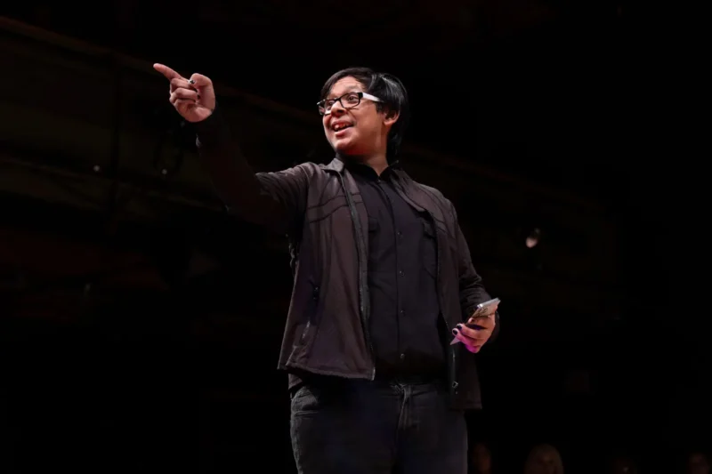 Person wearing glasses and dark clothing stands on stage, holding a phone and pointing with their right hand.