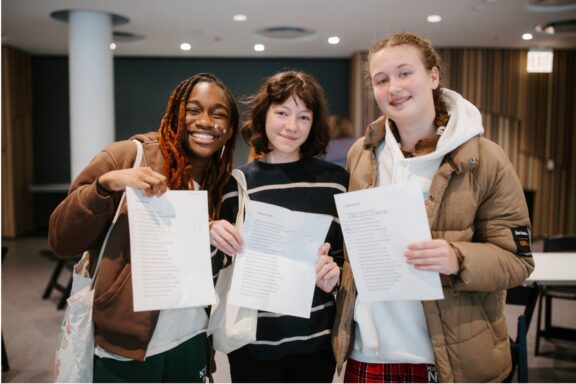 Three students standing indoors hold up printed documents and smile at the camera.