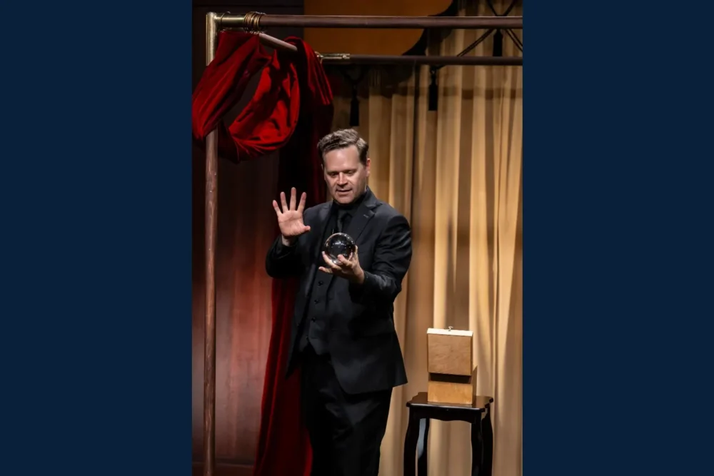 A man in a black suit stands on stage holding a clear crystal ball in one hand and raising the other hand, with a box on a small table beside him.