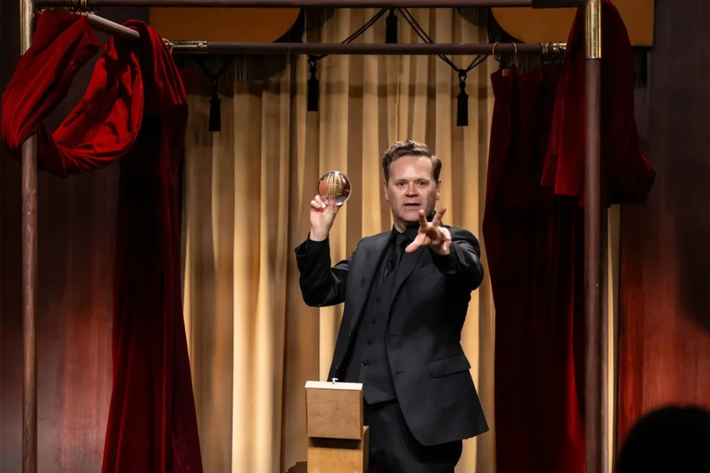 A man in a black suit stands on stage holding a clear sphere in one hand and gesturing with the other, in front of a curtain with red drapes.