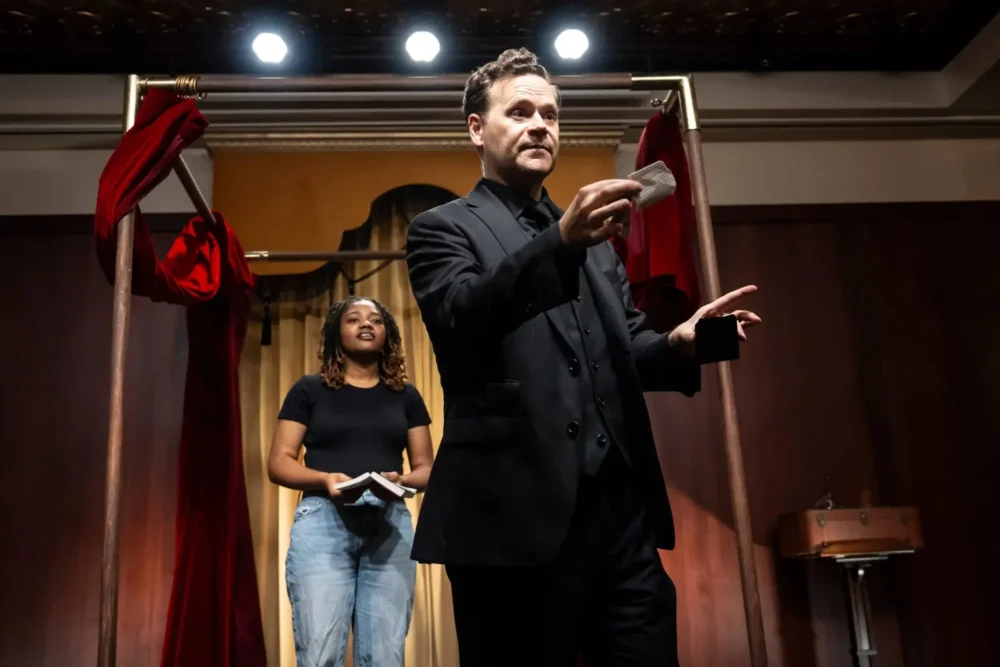 A man in black holds a playing card while performing on stage, with a woman in casual clothes standing behind him holding a notebook.