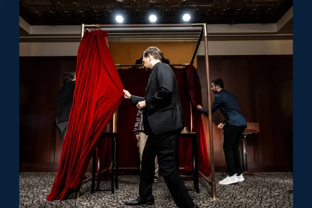 Four people interact with a booth framed by gold poles and red curtains in a carpeted, wood-paneled room under bright ceiling lights.