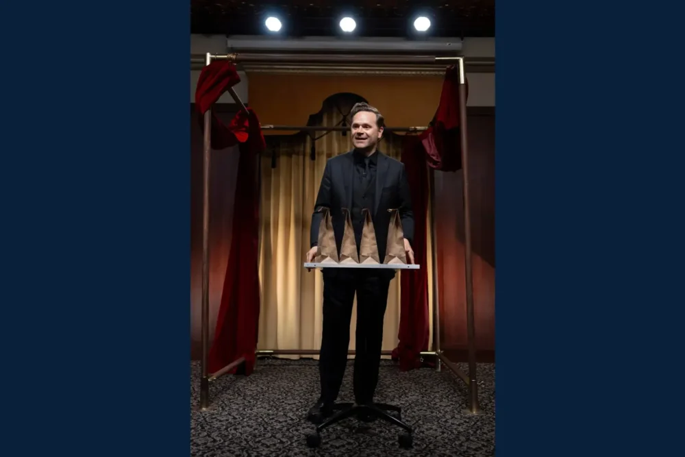 A man in a black suit stands on a patterned carpet holding a tray with four brown paper bags, with red curtains and stage lights in the background.