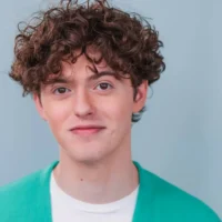 Young person with curly brown hair wearing a white shirt and teal jacket, standing against a plain light blue background and looking at the camera.