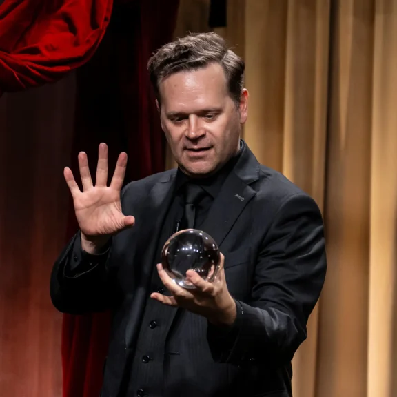 A man in a black suit holds a clear glass sphere in one hand and raises his other hand, standing in front of tan and red curtains.