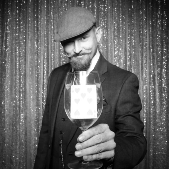 A man in a suit and flat cap holds a wine glass with a playing card inside, standing in front of a glittery curtain.