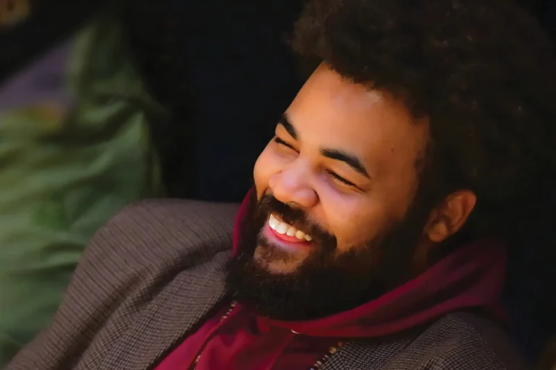A man with a beard and afro hair wearing a maroon hoodie and a brown blazer is smiling and looking slightly to the side.