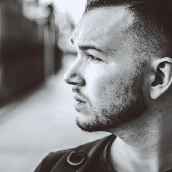 Black and white photo of a man with a trimmed beard looking to the left, wearing a backpack, with a blurred outdoor background.