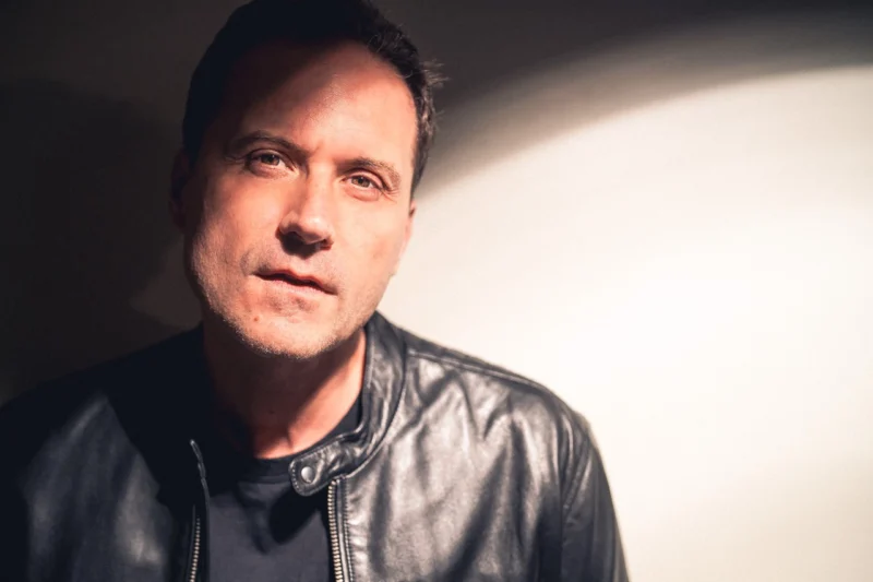 A man in a black leather jacket stands against a white background, looking directly at the camera, with light and shadow partially illuminating his face.