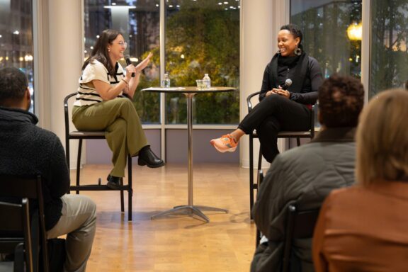 Two women sit on chairs at a small table in front of an audience, engaged in a discussion with microphones in hand, in a well-lit indoor space with large windows.