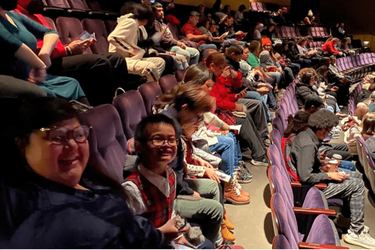 Audience members, including children and adults, sit in rows of purple seats inside a theater, waiting for a performance to begin.