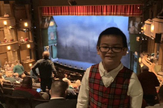 A boy wearing glasses and a plaid vest smiles in a theater with an audience and a stage set for a play in the background.