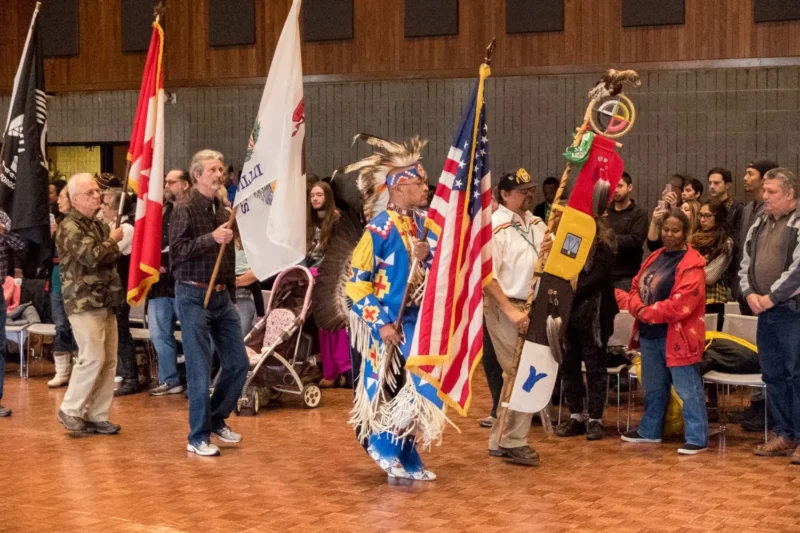 A group of people participate in an indoor event, holding flags and banners; one person in Indigenous regalia leads the procession as others stand and watch.