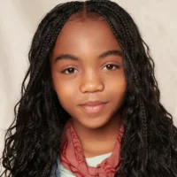 A young girl with long, wavy hair styled in braids, wearing a red scarf and light-colored top, poses in front of a neutral background.