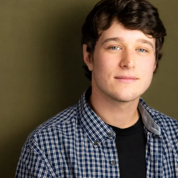 A young man with short brown hair, wearing a blue plaid shirt over a black t-shirt, poses in front of an olive green background.