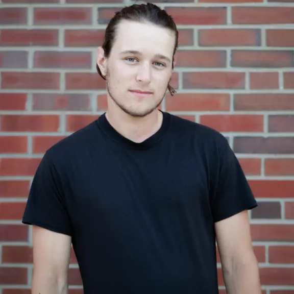A young man with light skin, brown hair pulled back, and blue eyes stands against a brick wall, wearing a plain black t-shirt.