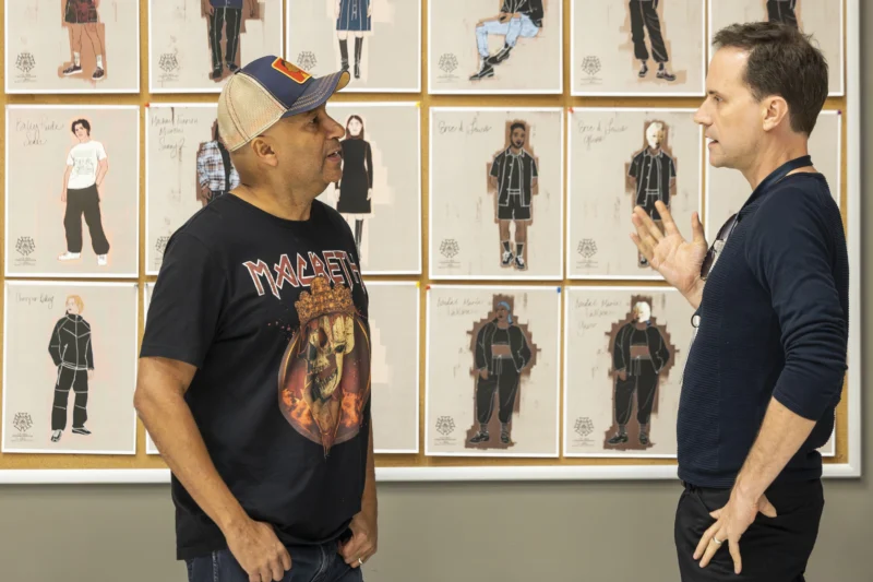 Two men talk in front of a wall covered with fashion sketches featuring different outfits and character designs.
