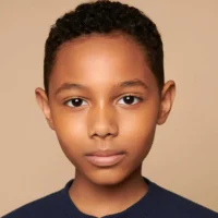 A young boy with short curly hair and brown skin looks directly at the camera with a neutral expression, against a plain beige background.