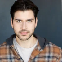 A man with dark hair, facial hair, and light-colored eyes wears a plaid shirt over a hoodie, facing the camera with a neutral expression against a plain dark background.