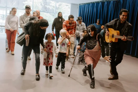 A group of adults and children walk indoors, led by a person in a skeleton costume and a musician playing a guitar. Blue curtains are in the background.