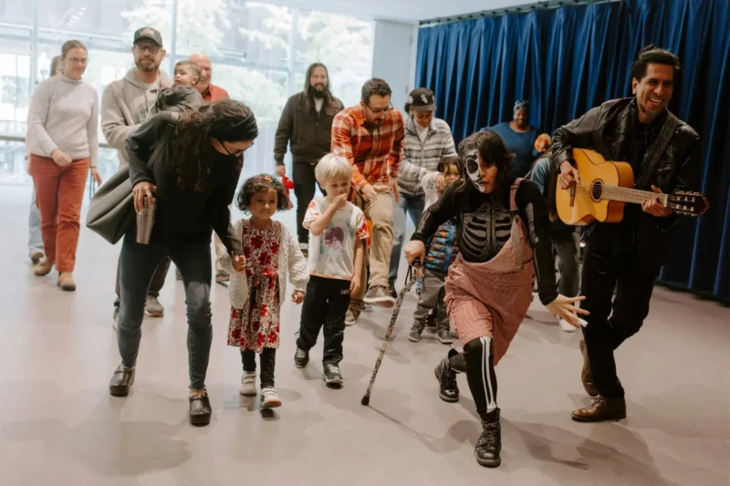 A group of adults and children walk indoors, led by a person in a skeleton costume and a musician playing a guitar. Blue curtains are in the background.