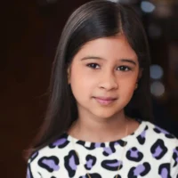 A young girl with long, dark hair wearing a white shirt with a purple and black animal print pattern.