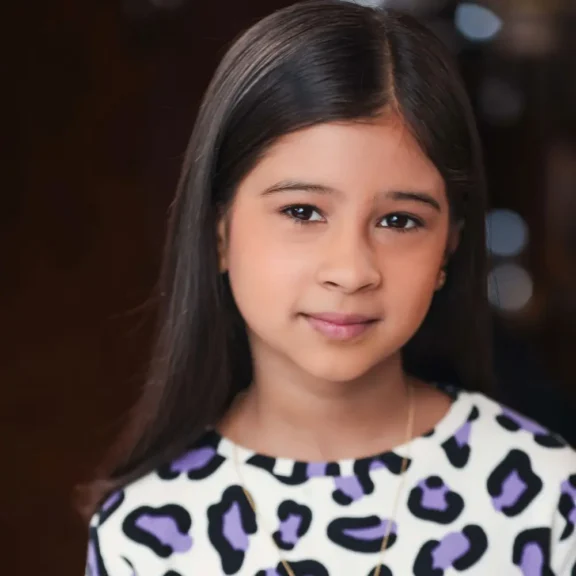 A young girl with long, dark hair wearing a white shirt with a purple and black animal print pattern.