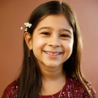 Young girl with long dark hair, smiling, wearing a red sequined top and small white flowers in her hair, standing in front of a plain brown background.