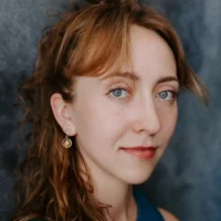A woman with light brown hair, blue eyes, and light skin looks at the camera. She wears a blue top and a single drop earring, with a neutral background behind her.