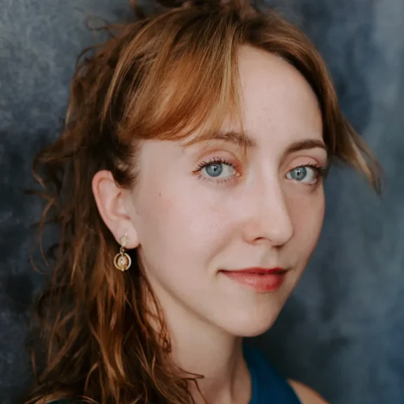 A woman with light brown hair, blue eyes, and light skin looks at the camera. She wears a blue top and a single drop earring, with a neutral background behind her.
