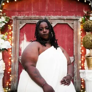 A person wearing a white dress sits in front of a rustic red wooden door decorated with flowers and string lights.