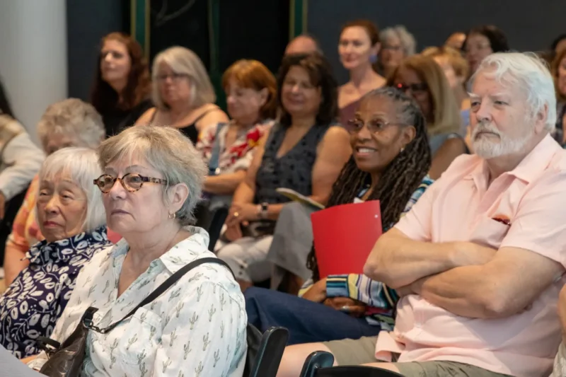 A diverse group of adults sits attentively in rows, listening to a speaker at an indoor event. Some hold papers or folders.
